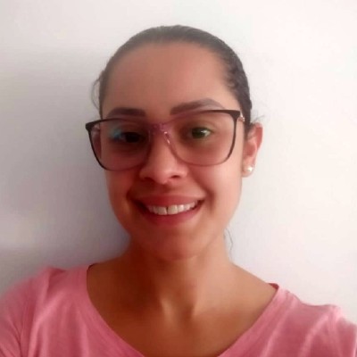 Foto de Juliana Cruz Gomes. De óculos redondos, cabelos presos em coque e camiseta cor de rosa, Juliana tem aparência bem jovem e sorri diante de uma parede branca